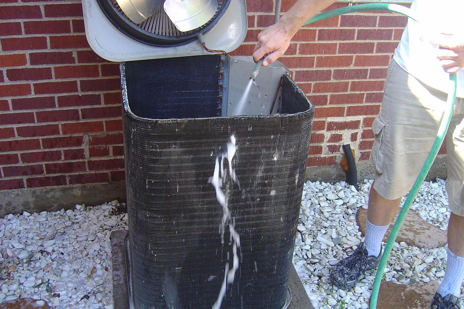 Garden hose rinsing the coil of an outdoor condenser next to a brick wall